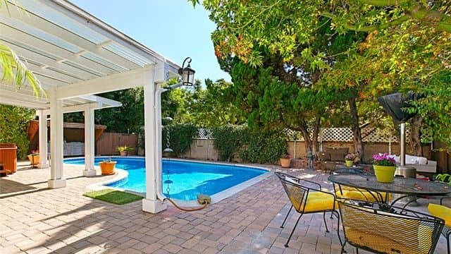 A backyard with a blue swimming pool, a paved patio, a white pergola, outdoor dining furniture with yellow cushions, and lush green trees and bushes. A hot tub is visible under the pergola on the left.